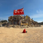 Kfar Khila herrian sarreran dagoen errotondako bandera gorri txiita. Atzean Hezbolako martirien argazkiak ageri dira deuseztutako etxe batean. Argazki hau atera zen unean biztanle eskas batzuk besterik ez zituen eta lapurretarik ez egoteko boluntarioak ziren. Argazkia: Andoni Lubaki / Ronak Press (Creative Commons By-Nd-Nc 4.0)