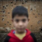 Mutiko baten erretratua Tel Nasri herrian, Yihadisten eta Turkiako Armadaren aurkako frontean, balaz estalitako horma baten aurrean. 2019ko urrian tropa estatubatuarrak Siria iparraldetik erretiratu ondoren, Turkiak erasoaldi bat hasi zuen herrialdearen ipar-ekialdeko eremu kurduen aurka. Gerra krimenak ikertzeko eskaerak mundu osotik iritsi ziren Hagako auzitegira baina ez zuten aintzakotzat hartu. Ankarak babestutako milizien jokabideari buruz nazioarteko medioek ezer gutxi esan dute. (Andoni Lubaki/Ronak Press)